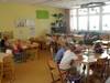 Das ist der Speiseraum. Dort stehen viele Tische und Stühle, da die Kinder hier ihr Mittagessen einnehmen. An einem Tisch sitzen mehrere Kinder und erledigen dort selbstständig ihre Hausaufgaben. In einem Regal sind Spielmaterialien, und an den Wänden hängen gestaltete Bilder von den Kindern. Im Hintergrund steht ein Klavier, auf dem die Kinder die Möglichkeit zum Üben haben.